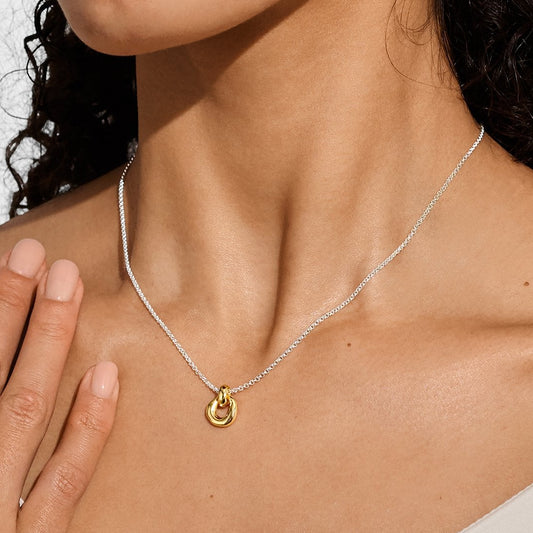 A close-up of a model wearing a delicate silver chain necklace with a small gold interlocking rings pendant, paired with a white top.