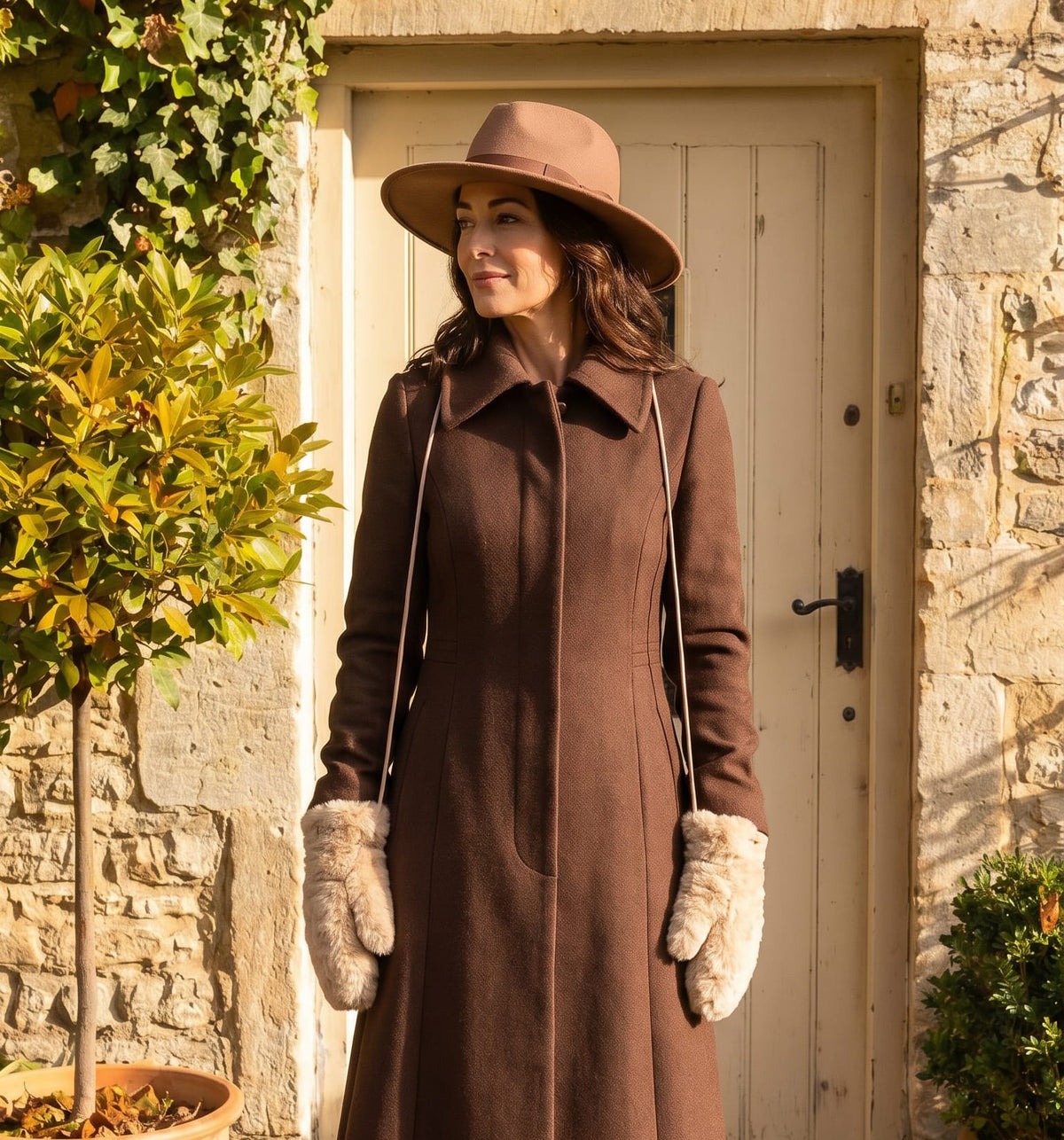 A woman stands outdoors in front of a stone wall and wooden door wearing a long brown coat, wide-brim brown hat, cream gloves, and black boots, with fallen leaves on the ground.