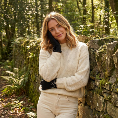 A woman stands outdoors in a wooded setting next to a stone wall, wearing a cream ribbed knit sweater and matching cream trousers with black gloves.