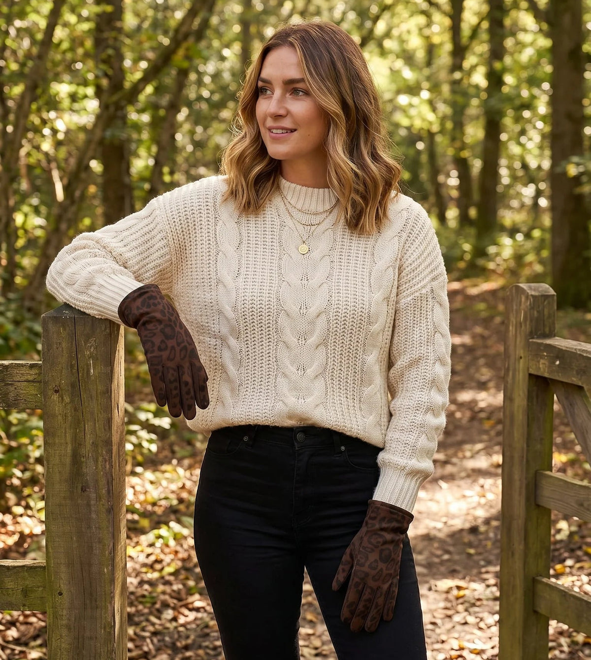 A woman wearing a cream cable-knit sweater, black pants, and dark brown gloves stands in a wooded outdoor setting next to a wooden gate.
