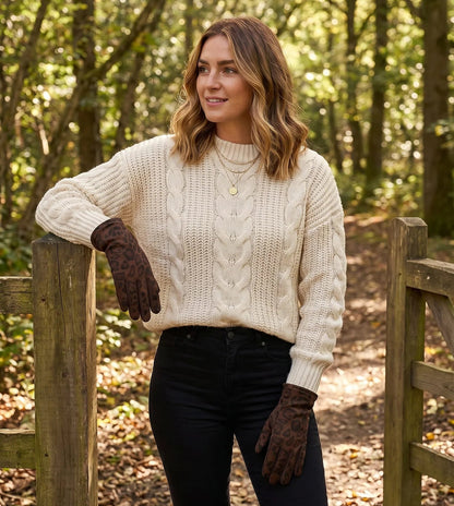 A woman wearing a cream cable-knit sweater, black pants, and dark brown gloves stands in a wooded outdoor setting next to a wooden gate.