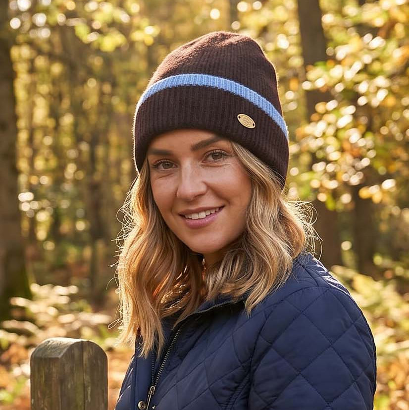 A woman wearing a navy quilted jacket and a brown beanie with a light blue stripe stands outdoors in a wooded setting with autumn foliage.