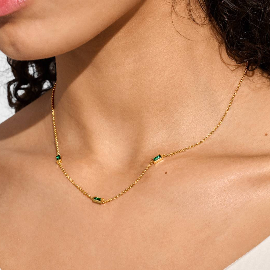 A close-up of a model wearing a delicate gold chain necklace with three small green rectangular stone accents, styled with a white top against a clean white background.