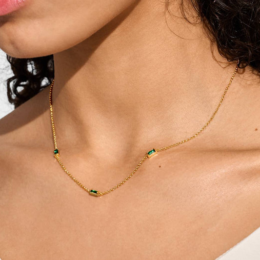 A close-up of a model wearing a delicate gold chain necklace with three small green rectangular stone accents, styled with a white top against a clean white background.